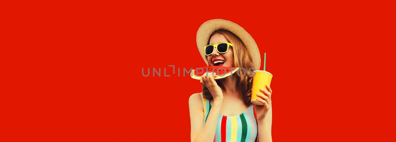 Summer portrait of happy smiling young woman eating fresh juicy slice of watermelon with cup of juice wearing tourist straw hat on red background