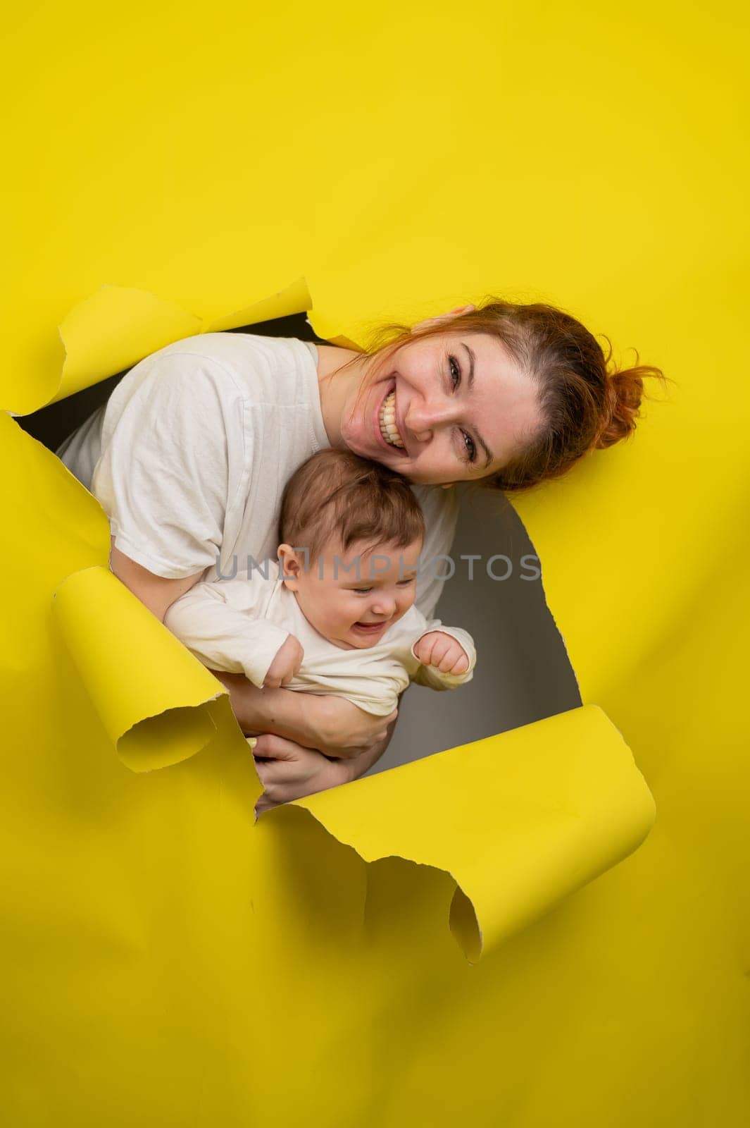 Cheerful Caucasian woman with little son rips and leans out through yellow cardboard background. Vertical photo. by mrwed54