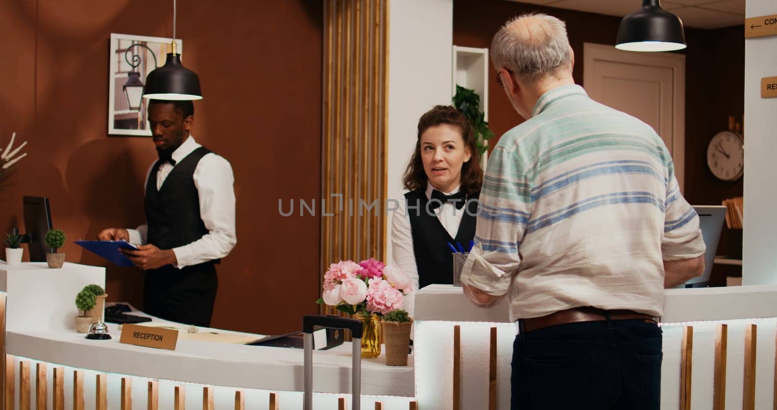 Elderly person arriving at hotel front desk with trolley bags, asking for assistance to check in. Receptionist ensuring pleasant stay for old man at all inclusive holiday retreat, tourism industry.