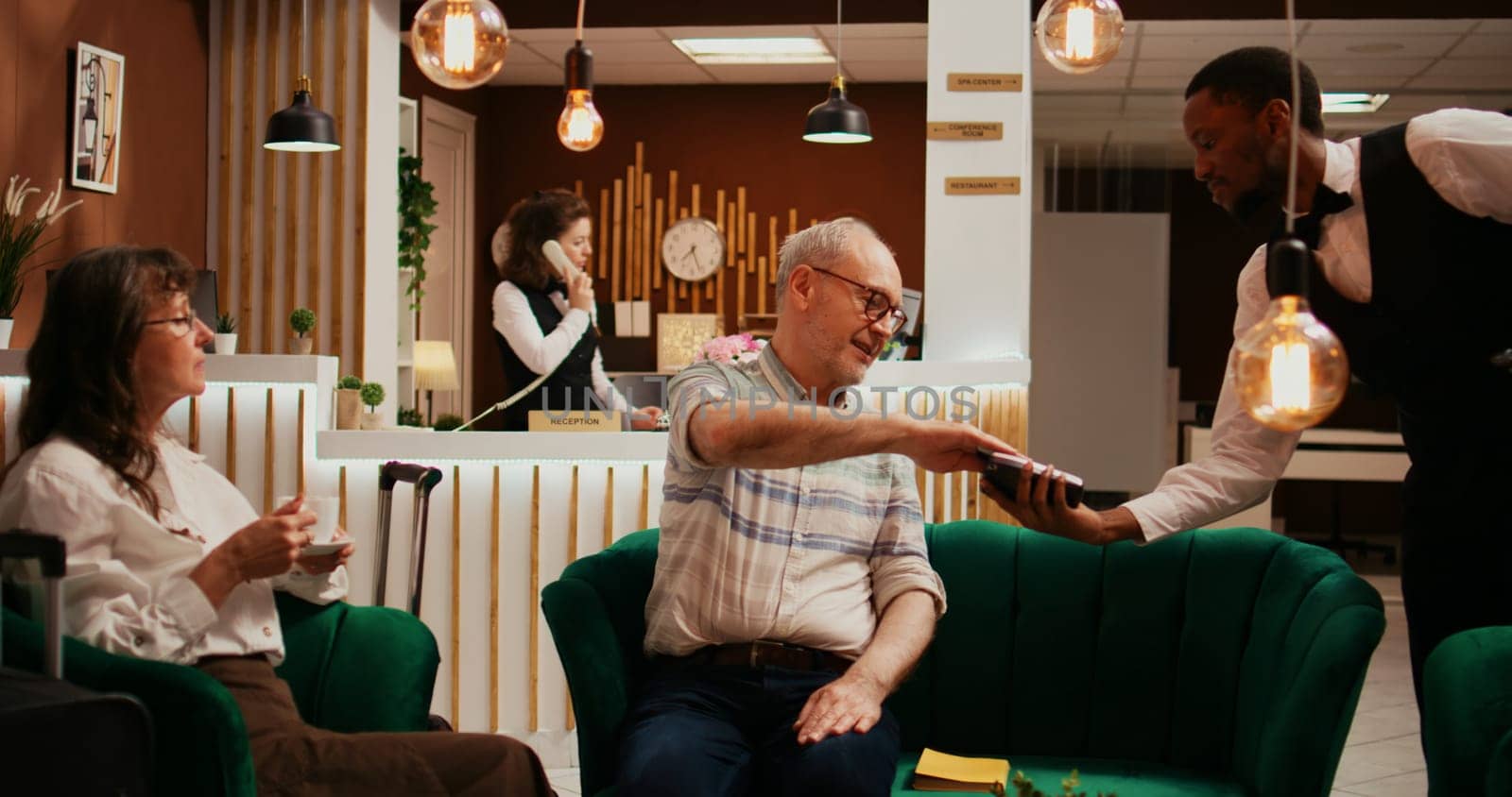 Retired couple paying for coffee cup from hotel bar, taking out credit card for pos terminal transaction. Husband and wife relaxing together in lounge area at resort, paying for services.