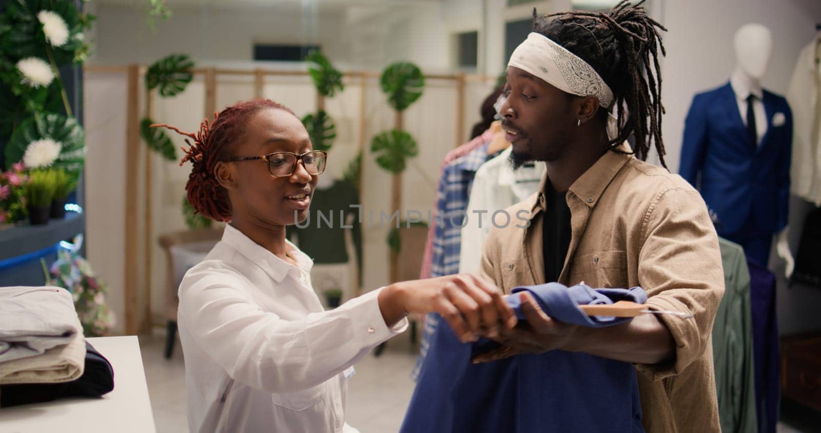 Employee assisting customer in luxury fashion boutique, helping him try on elegant clothes. African american worker handing client suiting high class men blazer in trendy clothing store
