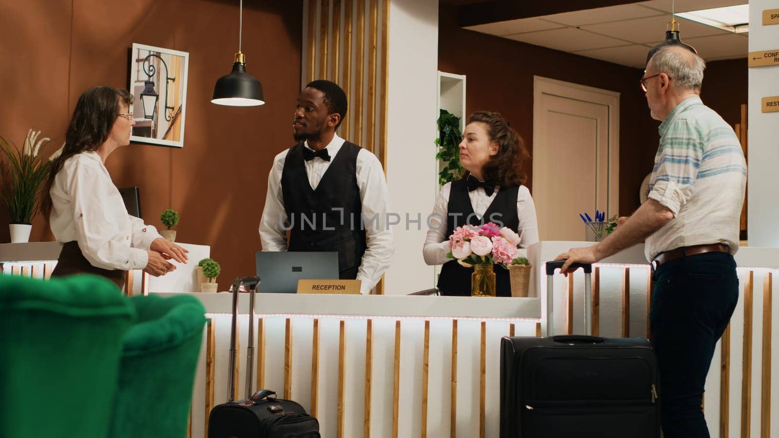 After check in verification, retired people receive room access with key card at hotel. Diverse employees assisting older couple with luggage, providing luxurious concierge services. Handheld shot.