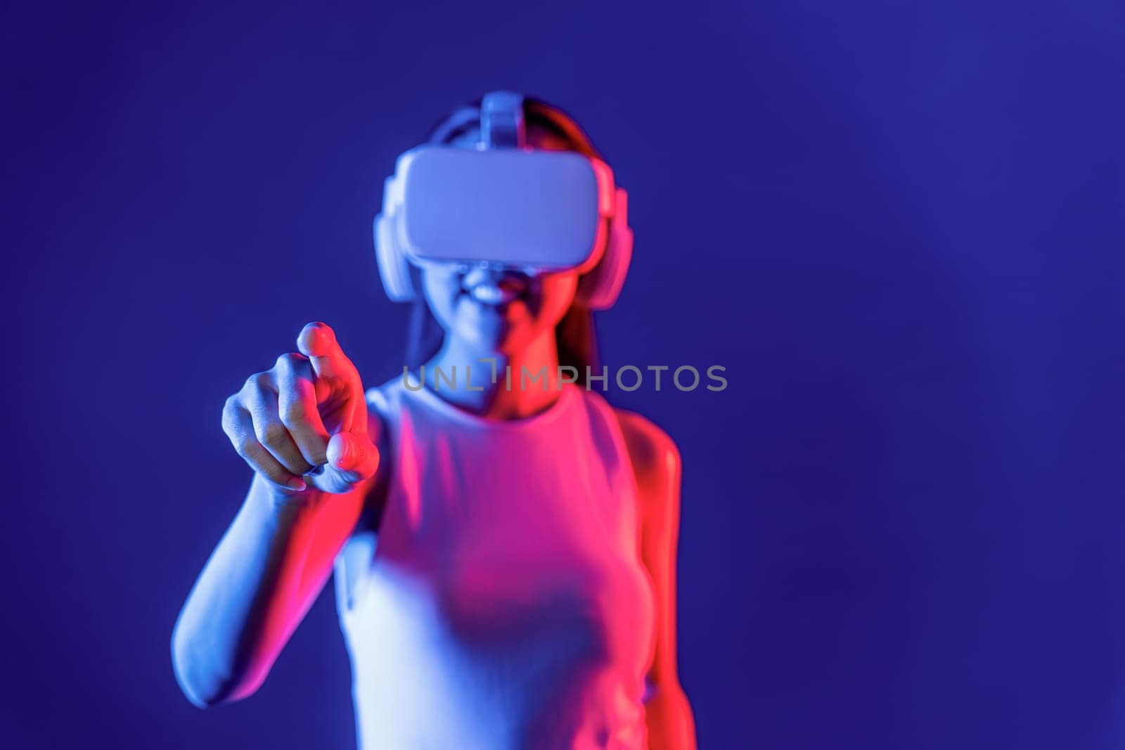 Smart female stand with surrounded by cyberpunk neon light wear VR headset connecting metaverse, futuristic cyberspace community technology. Woman using finger pointing virtual object. Hallucination.