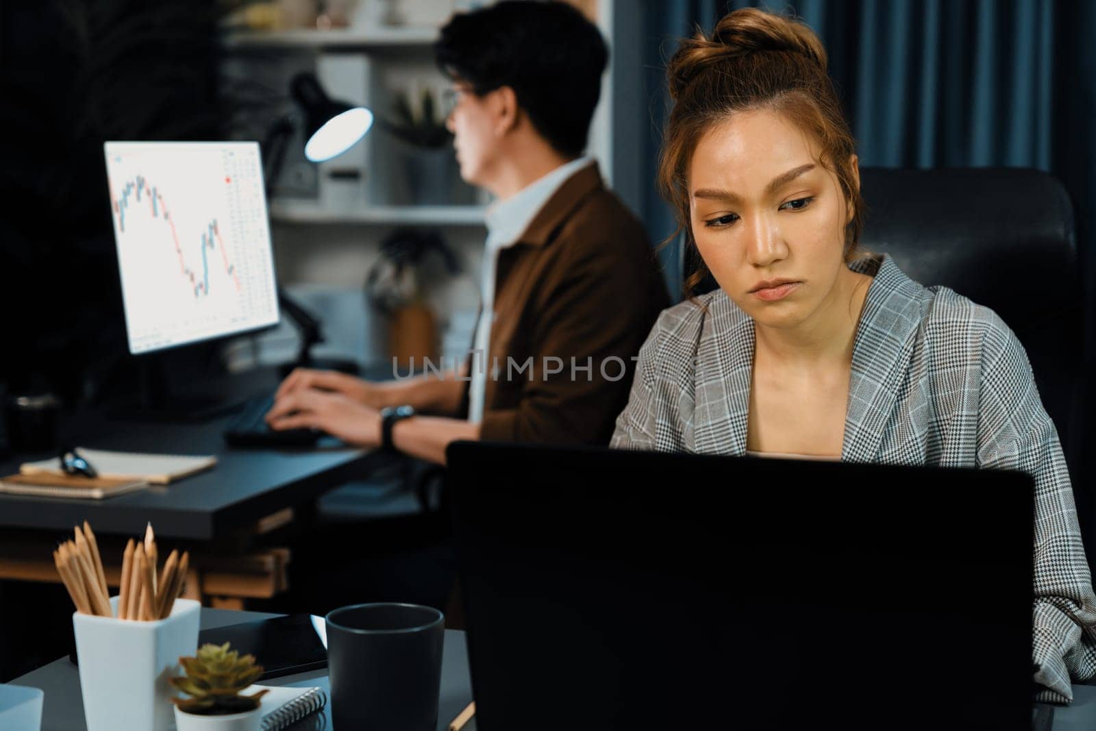 Serious face of thinking businesswoman focusing computer to report data analysis social media online project job with coworker working at back side at over late night time at modern office. Infobahn.