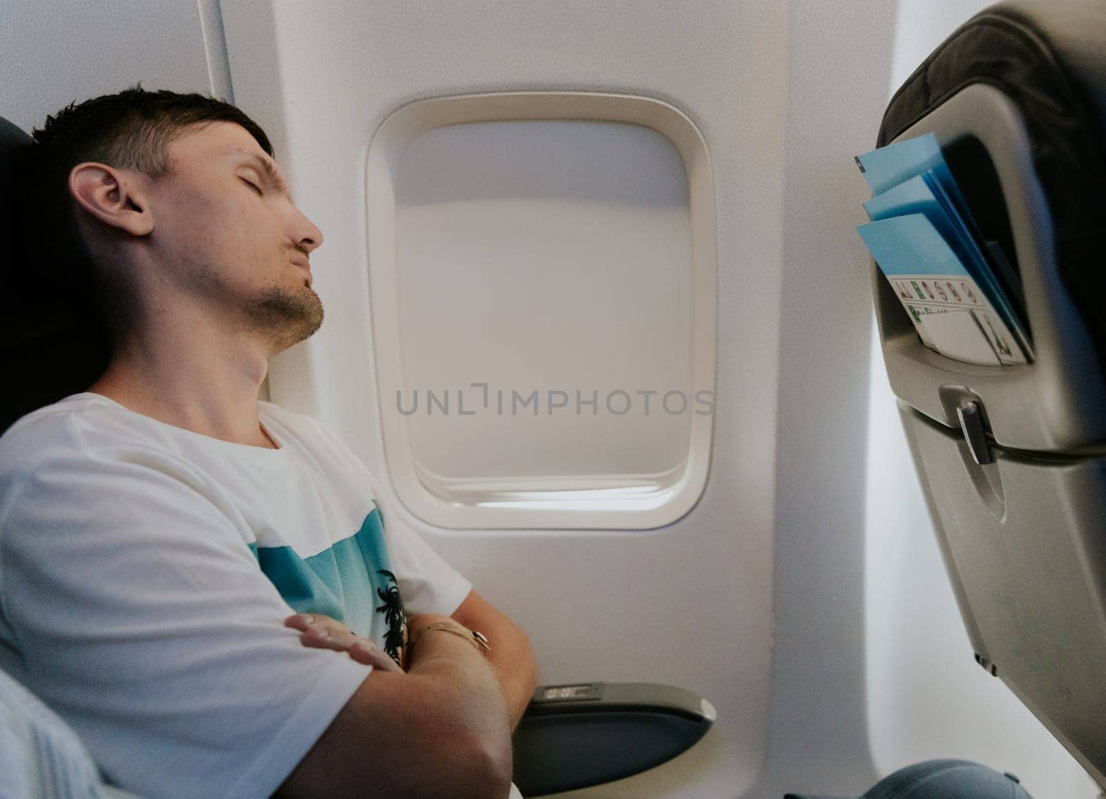 One young handsome Caucasian guy sleeps sweetly during a flight while sitting with his arms crossed near a closed curtain window in an airplane on a summer day, close-up side view.