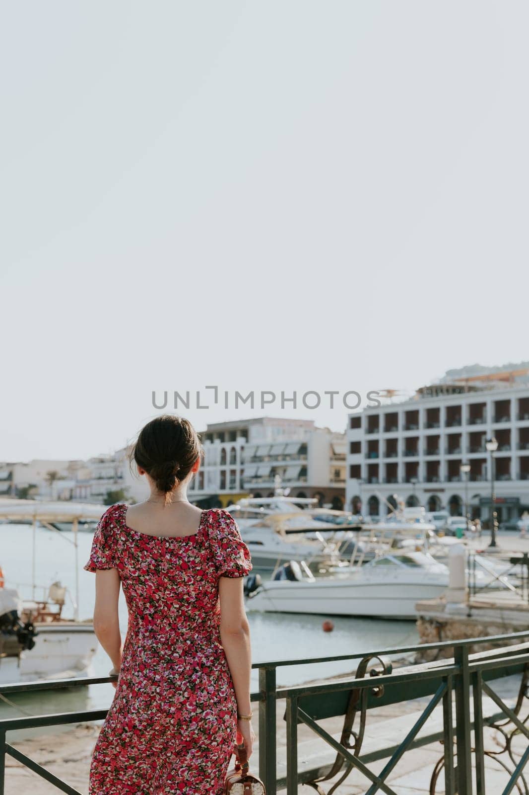 A portrait of one young beautiful Caucasian brunette woman in a bright colorful dress stands with her back to the left, leaning against an iron railing against the backdrop of a blurred embankment with boats on a sunny summer day, close-up side view.