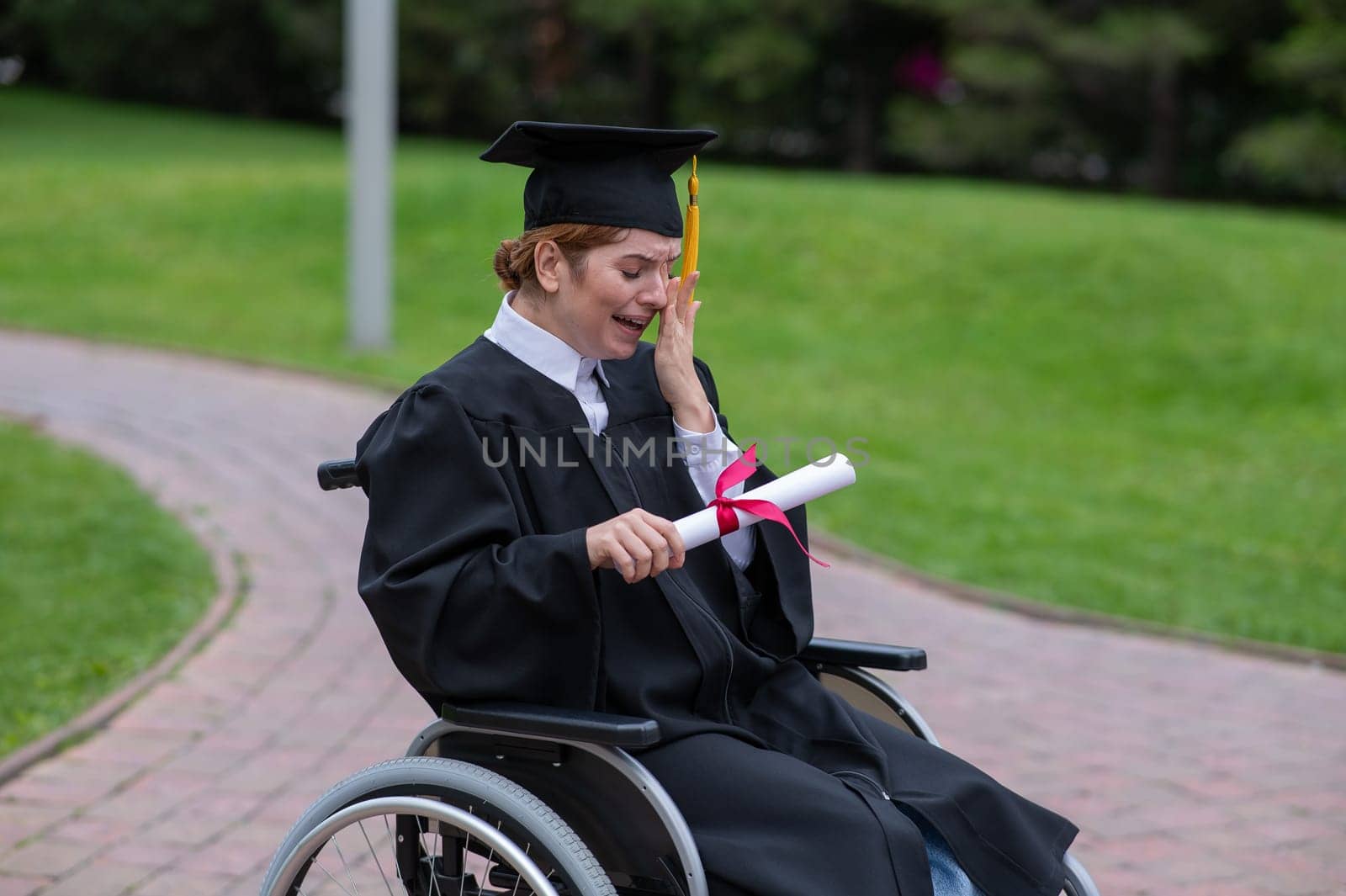 A caucasian woman in a wheelchair holds her diploma and cries with joy outdoors. by mrwed54