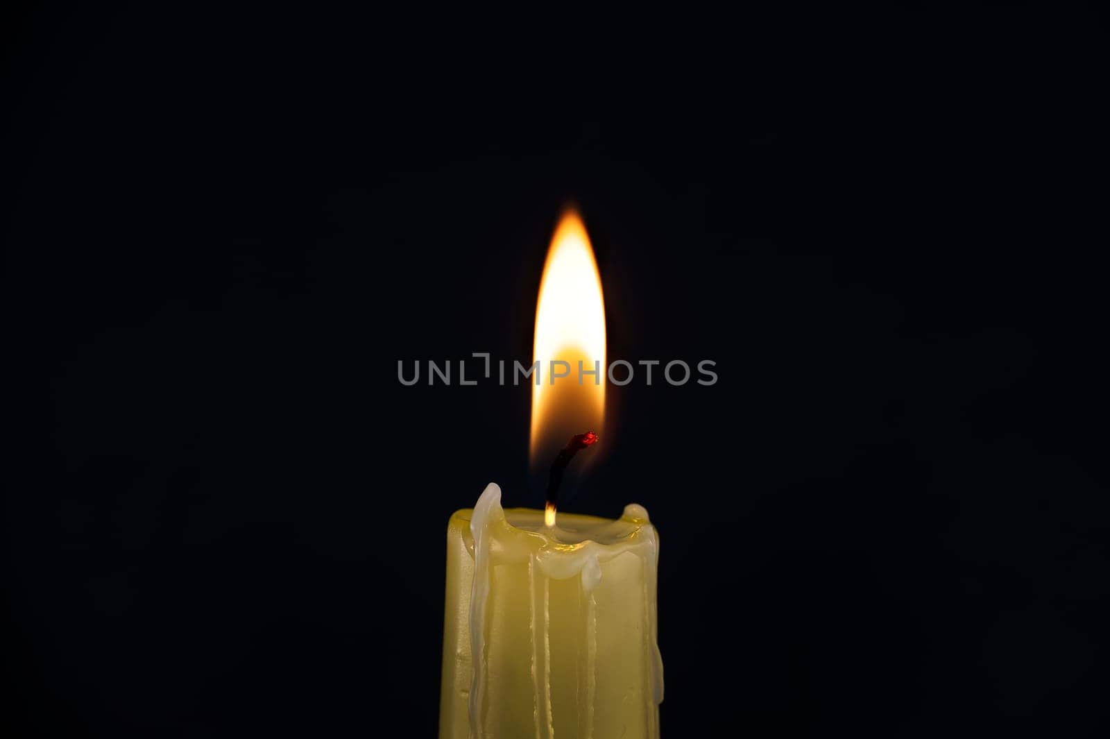 Burning yellow wax candle contrasting with the dark background, candle's gentle flame suggesting of remembrance, reflection and hope