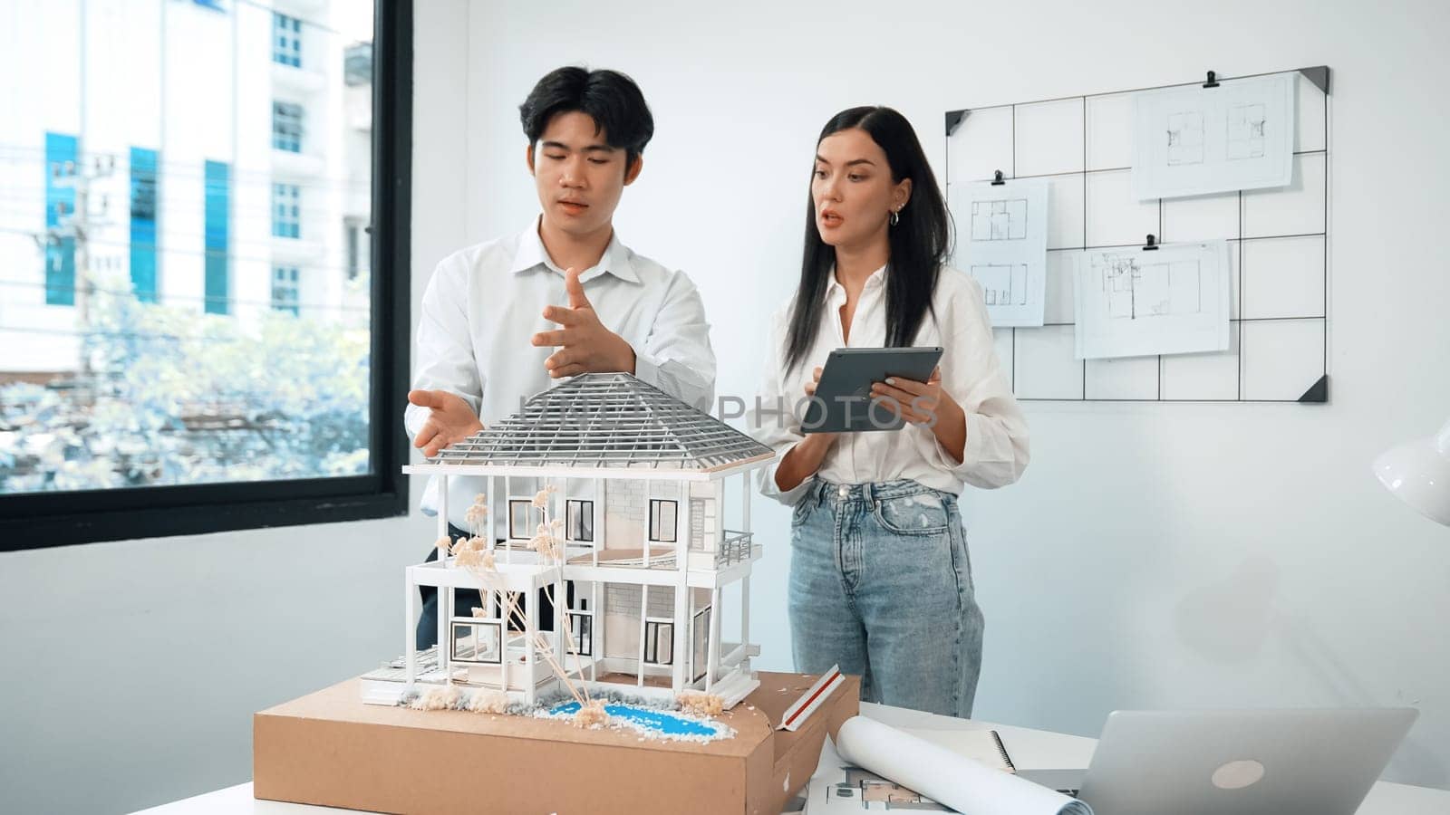 Beautiful young caucasian project manager tells asian engineer about building detail by using tablet while engineer measuring house model on meeting table with blueprint scatter around. Immaculate