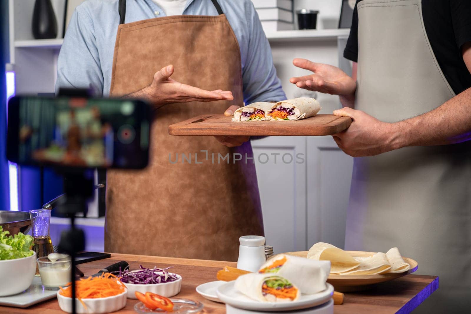 Cropped view of chef influencers presenting fresh salad roll on cooking step by step, streaming via smartphone on social media live channel, showing ingredients vegetable soft tortilla wrap. Sellable.