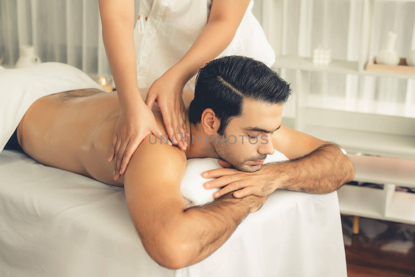 Caucasian man customer enjoying relaxing anti-stress spa massage and pampering with beauty skin recreation leisure in day light ambient salon spa at luxury resort or hotel. Quiescent