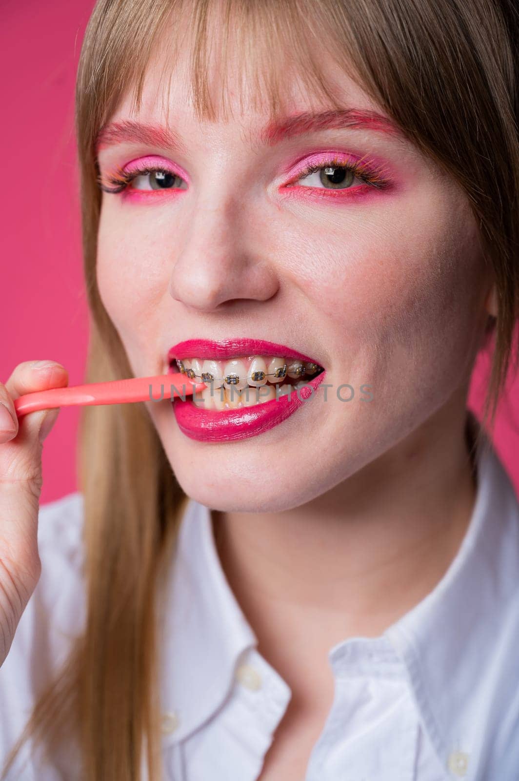 Portrait of a young woman with braces and bright makeup chewing gum on a pink background. Vertical photo