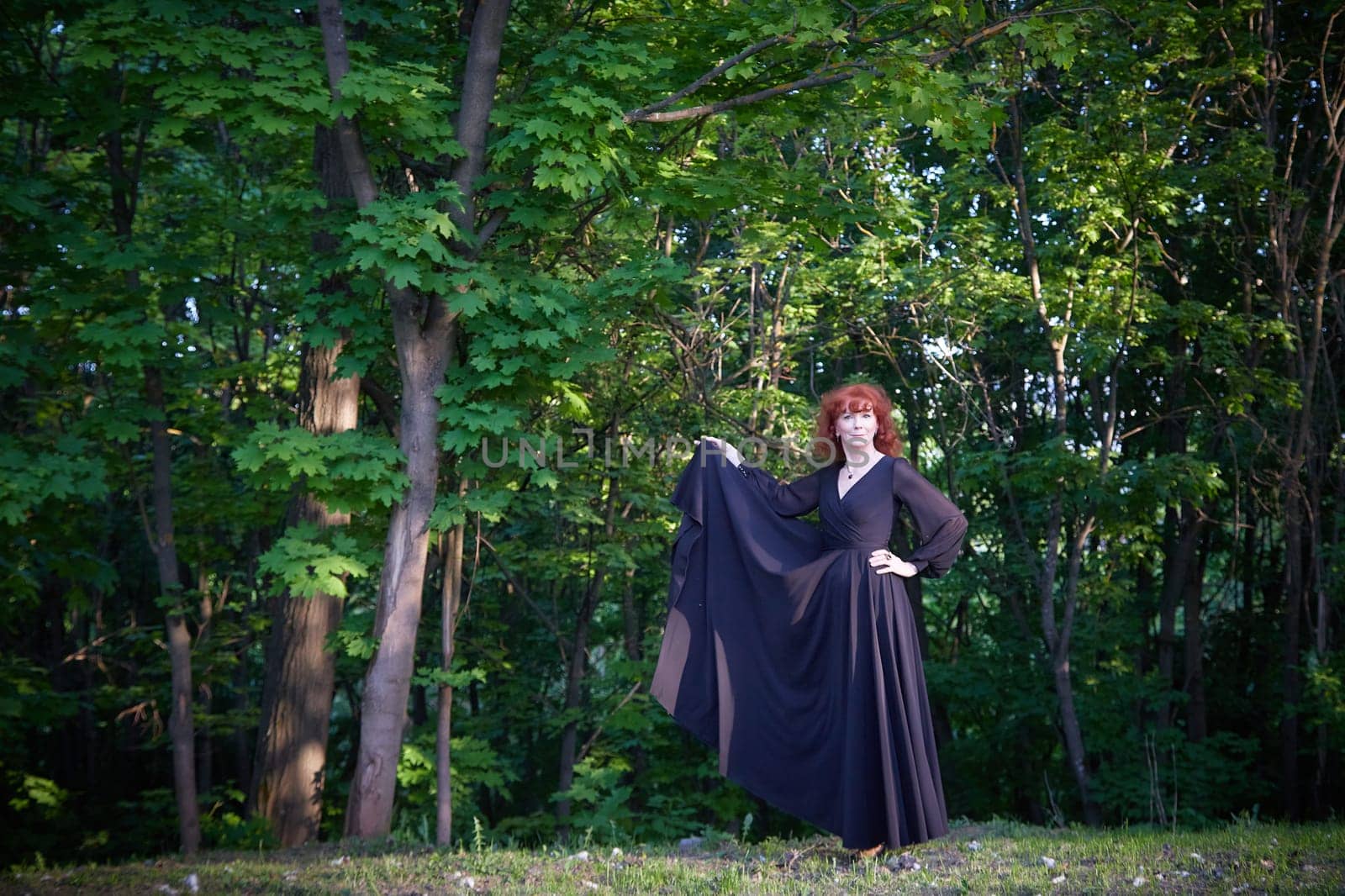 Elegant senior mature Woman dancing in Black Dress in green park. Woman with red hair posing on nature