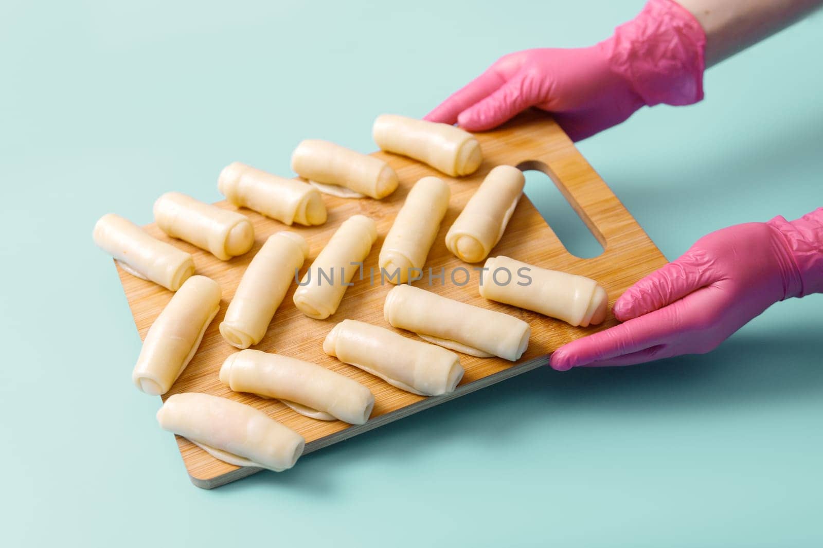 The hands of the pastry chef hold a wooden board on which raw sweet dough rolls lie neatly on a blue background, frozen semi-finished products.