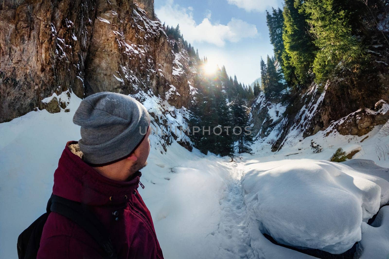 A man wearing a hat stands in the freezing snow, admiring the sun over a mountainous landscape. Snowcovered trees and clouds fill the sky. Almaty nature, Central asia, Kazakhstan