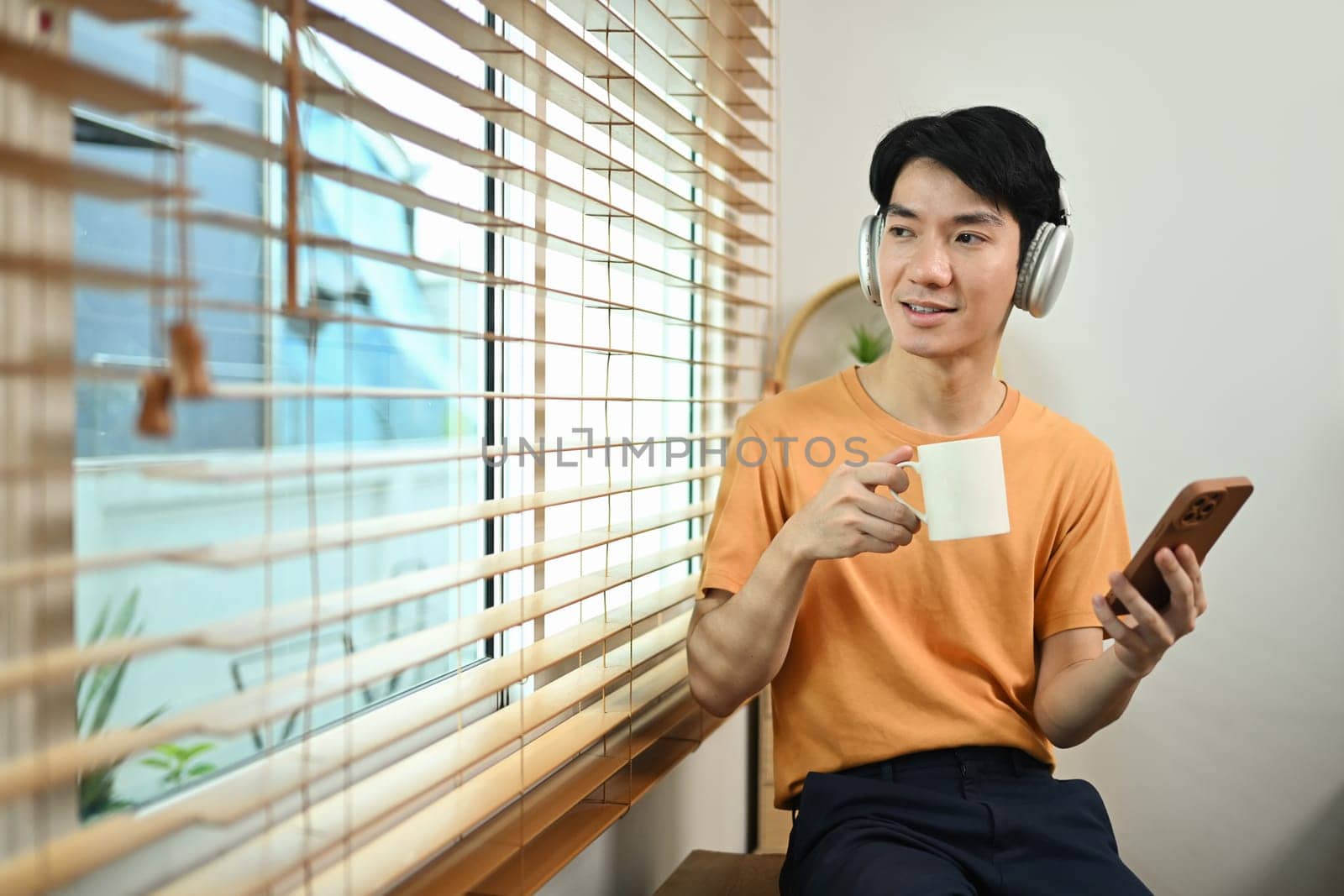 Carefree young asian man sitting by the window with a cup of coffee and looking away.