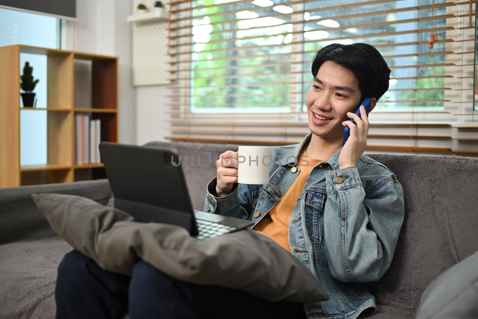 Smiling young man having phone conversation while drinking coffee on couch at home.