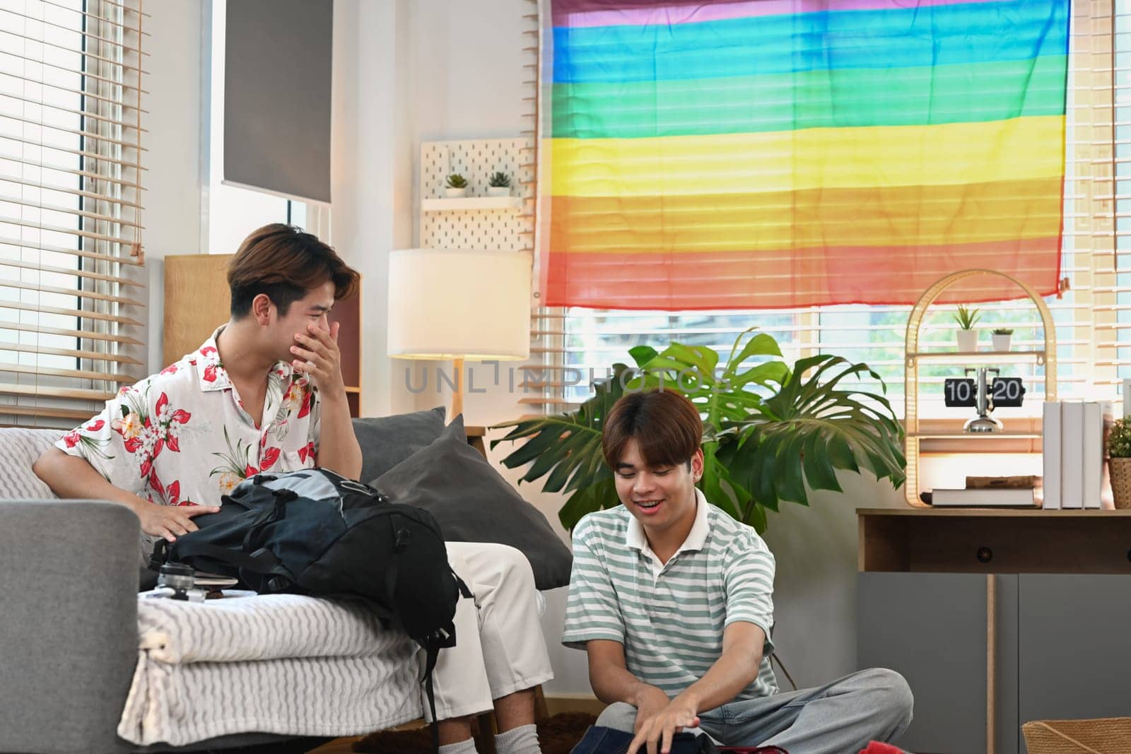 Happy young gay couple packing suitcase on floor in living room with rainbow flag.