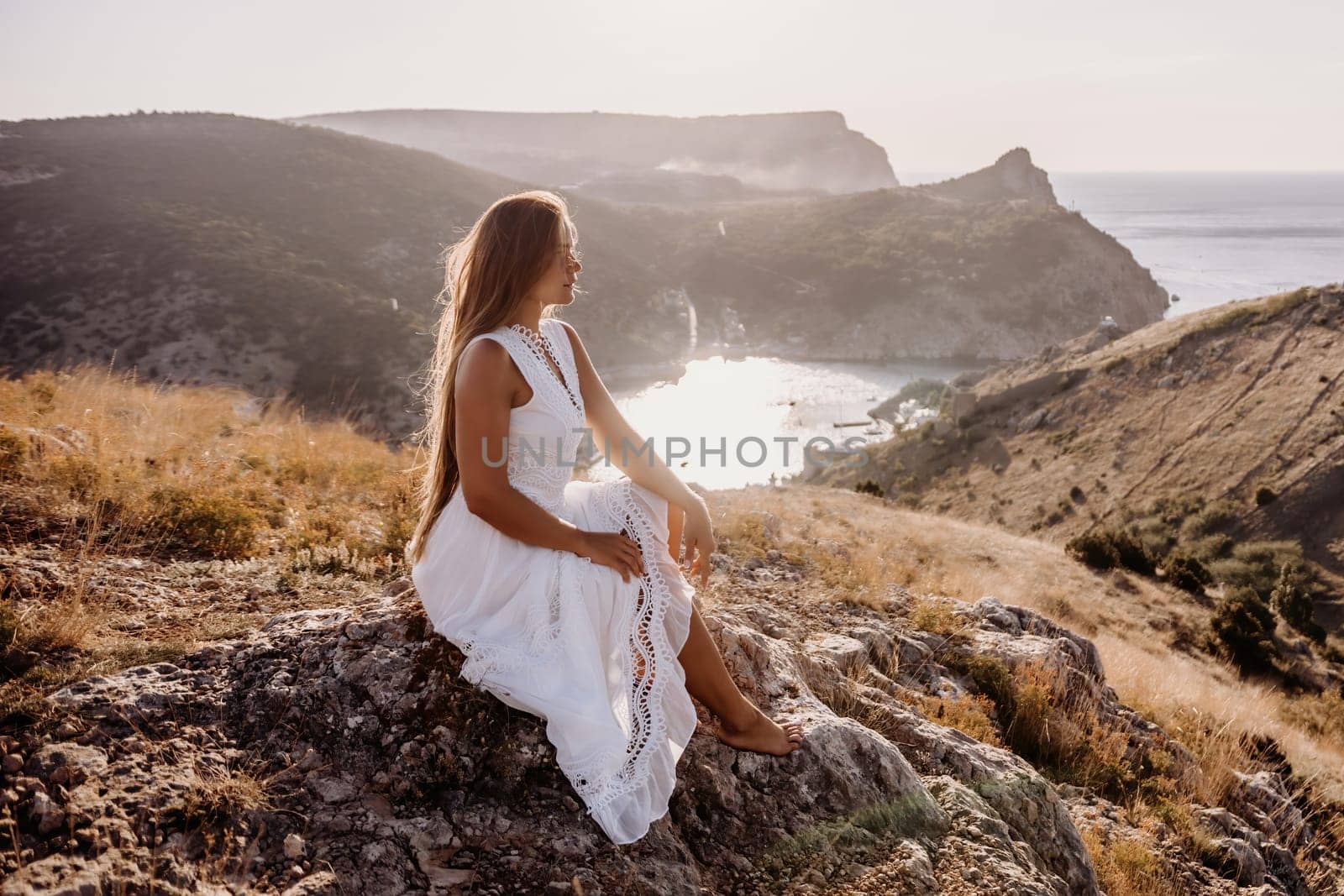 A woman in a white dress sits on a rock overlooking a body of water. The scene is serene and peaceful, with the woman enjoying the view and the calmness of the surroundings