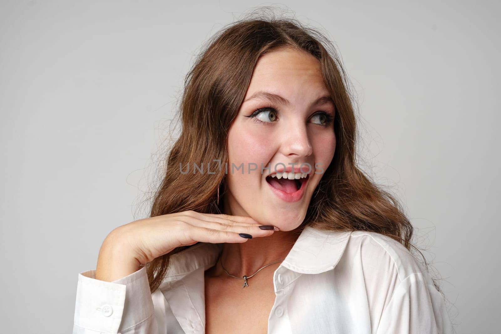 A young woman with long brown hair and a white blouse exhibits a look of joyful surprise, placing one hand delicately under her chin while her eyes gaze to the side with a bright, expressive smile.