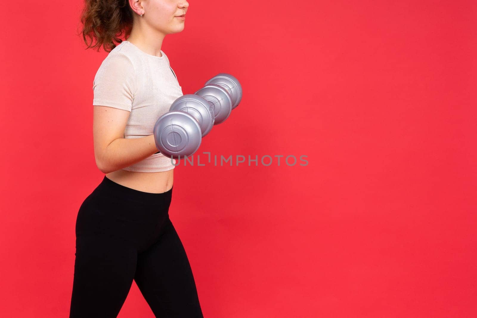 Teenage sportive girl exercises with dumbbells to develop muscles isolated on red background. by Zelenin