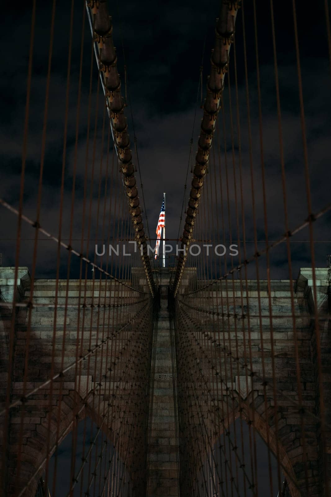 A captivating view of the Brooklyn Bridge at night featuring an American flag illuminated at the center. The iconic structure is showcased against a dark sky, highlighting its architectural beauty.