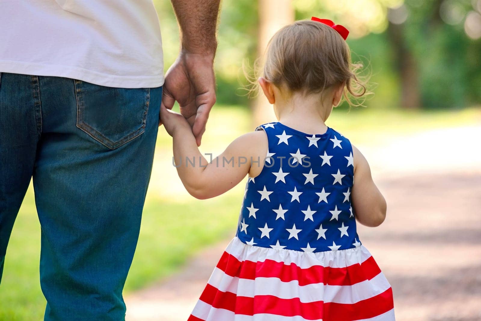 Child in a flag dress holds an adults hand while walking outdoors on Independence Day.