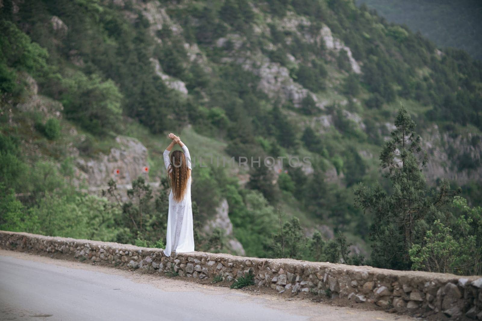A woman stands on a stone wall overlooking a mountain. She is wearing a white dress and she is in a contemplative mood