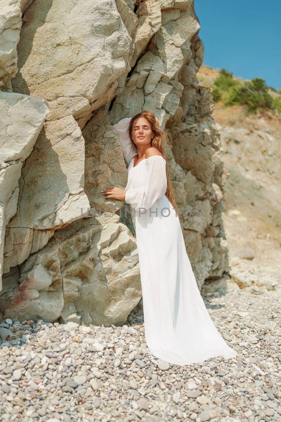 A woman in a white dress is standing on a rocky beach. Concept of serenity and tranquility, as the woman is enjoying the natural beauty of the beach. by Matiunina