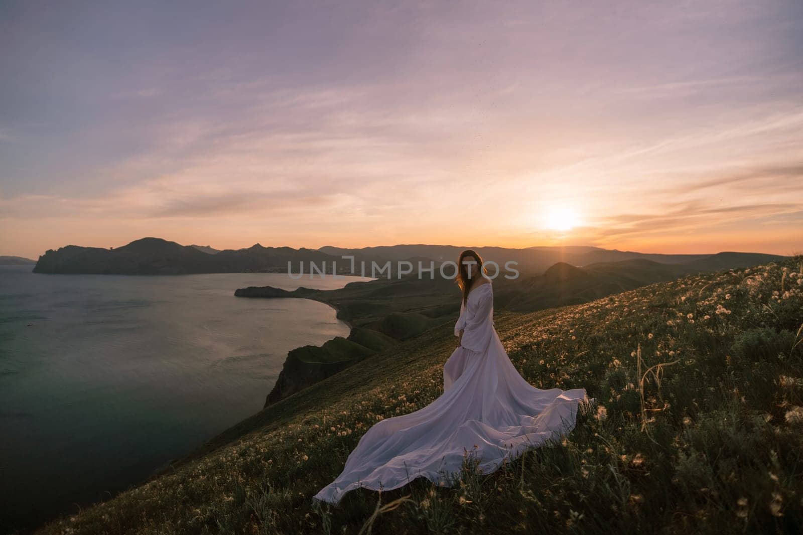 A woman stands on a hill overlooking a body of water. The sky is a mix of blue and orange, and the sun is setting. The woman is wearing a long white dress, and she is enjoying the view