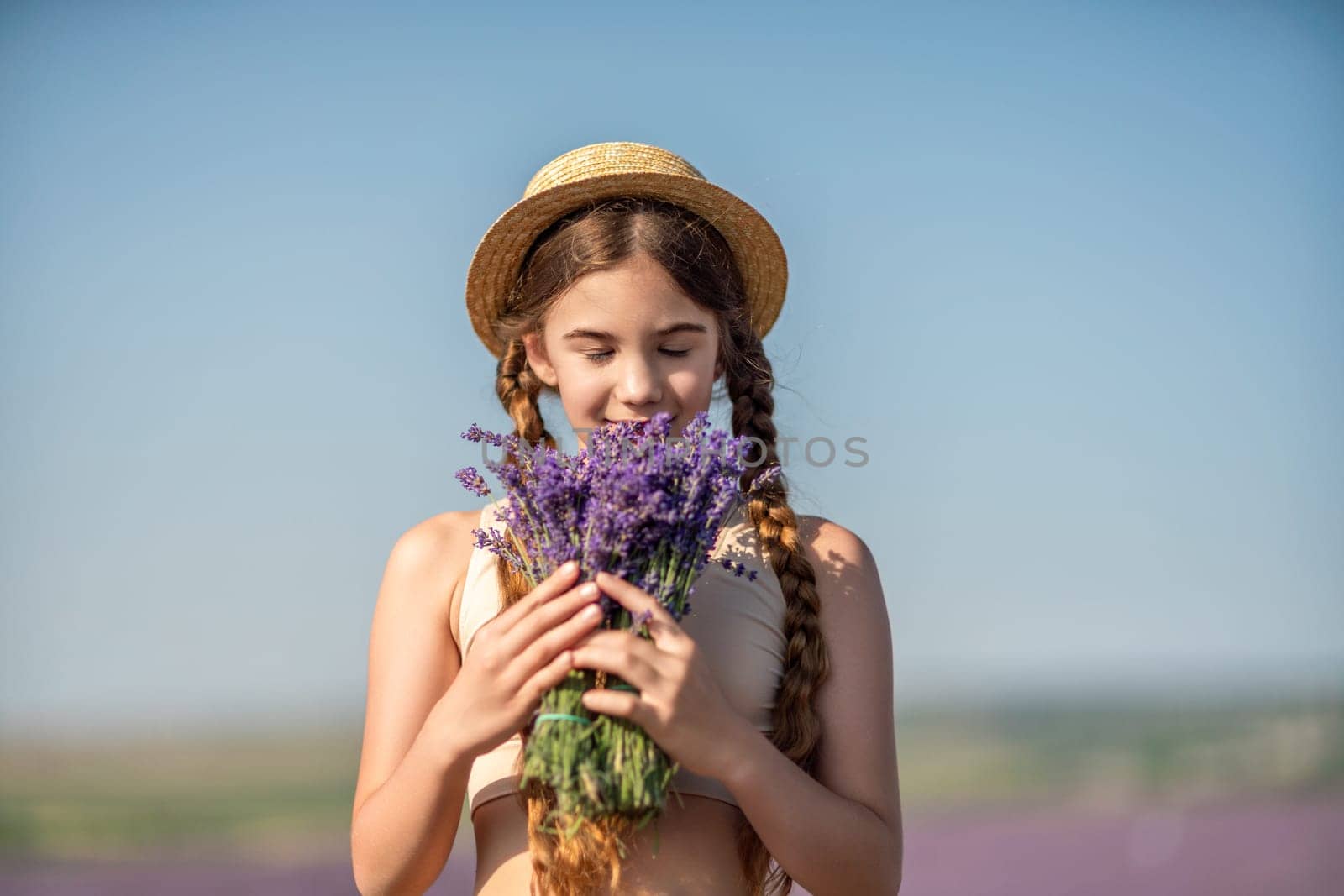 girl is holding a bunch of lavender purple flowers in her hands and wearing a straw hat. She is smiling and she is enjoying herself. The scene is set in a field of lavender, which adds to the peaceful. by Matiunina