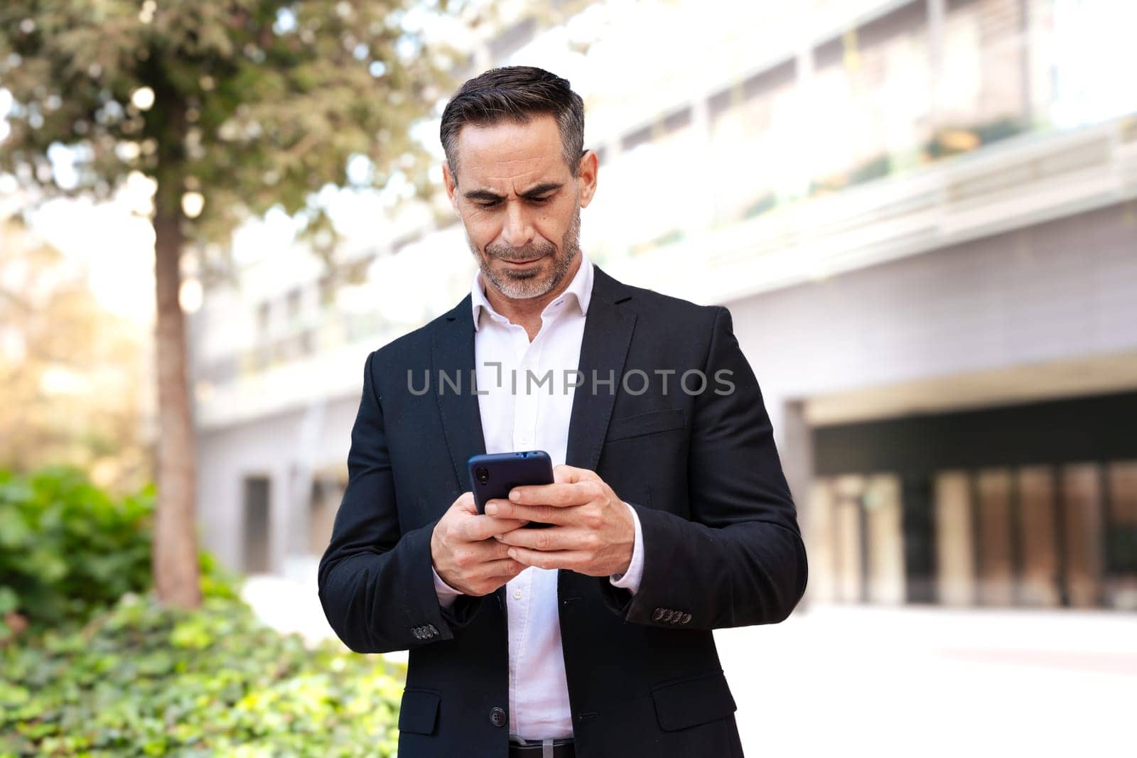 Satisfied with the results, the businessman walks down the street outside the office building, a mature boss holds a phone and coffee in his hands, writes messages and reads news online, using an app.