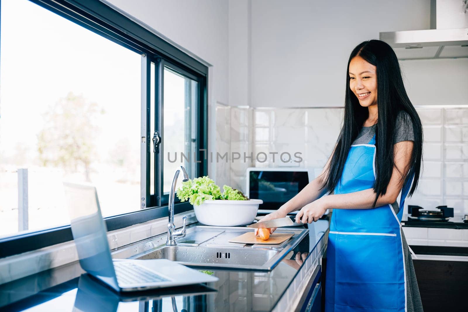 A woman stands in the kitchen enjoying cooking while searching for tutorials on her laptop by Sorapop