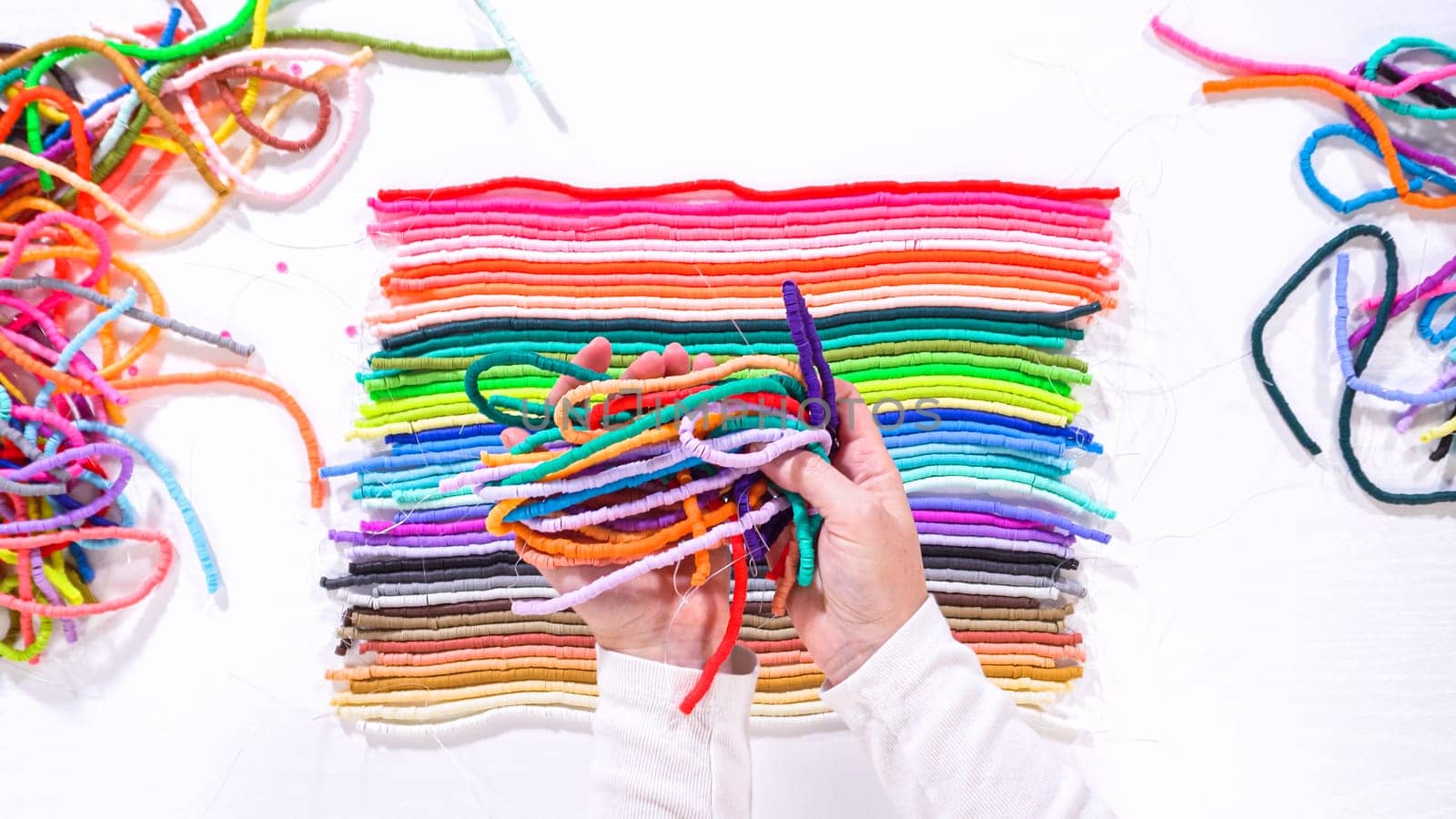 Flat lay. Artistic hands lift a selection of brightly colored clay bead cords, contrasting with organized layers beneath on a white background.