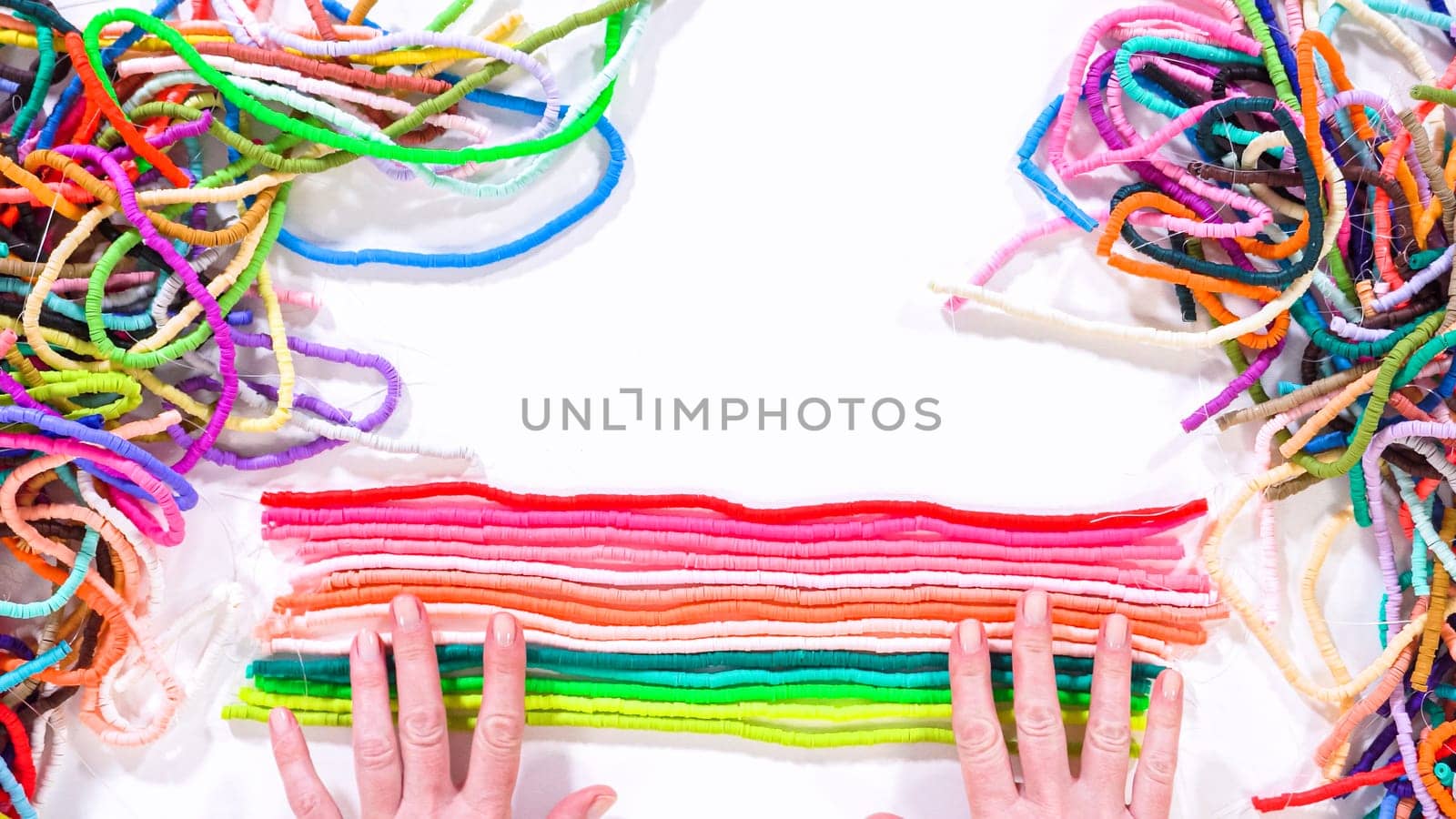 Flat lay. Hands touch a neatly organized rainbow of colorful, coiled clay bead strands against a contrasting white surface.