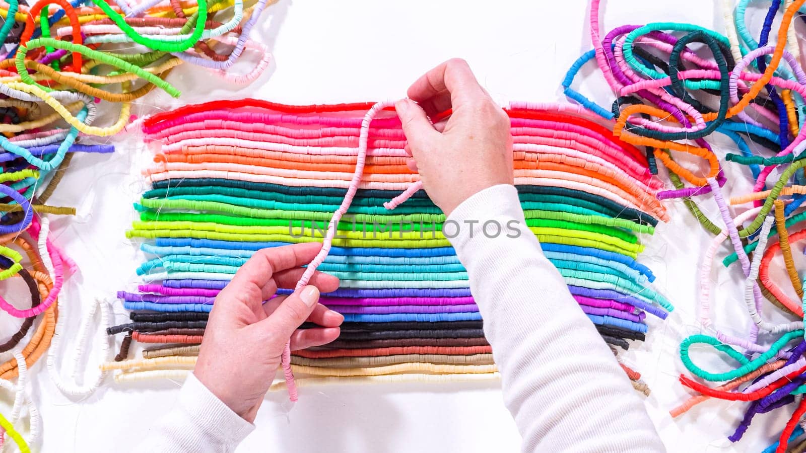 Flat lay. Hands touch a neatly organized rainbow of colorful, coiled clay bead strands against a contrasting white surface.