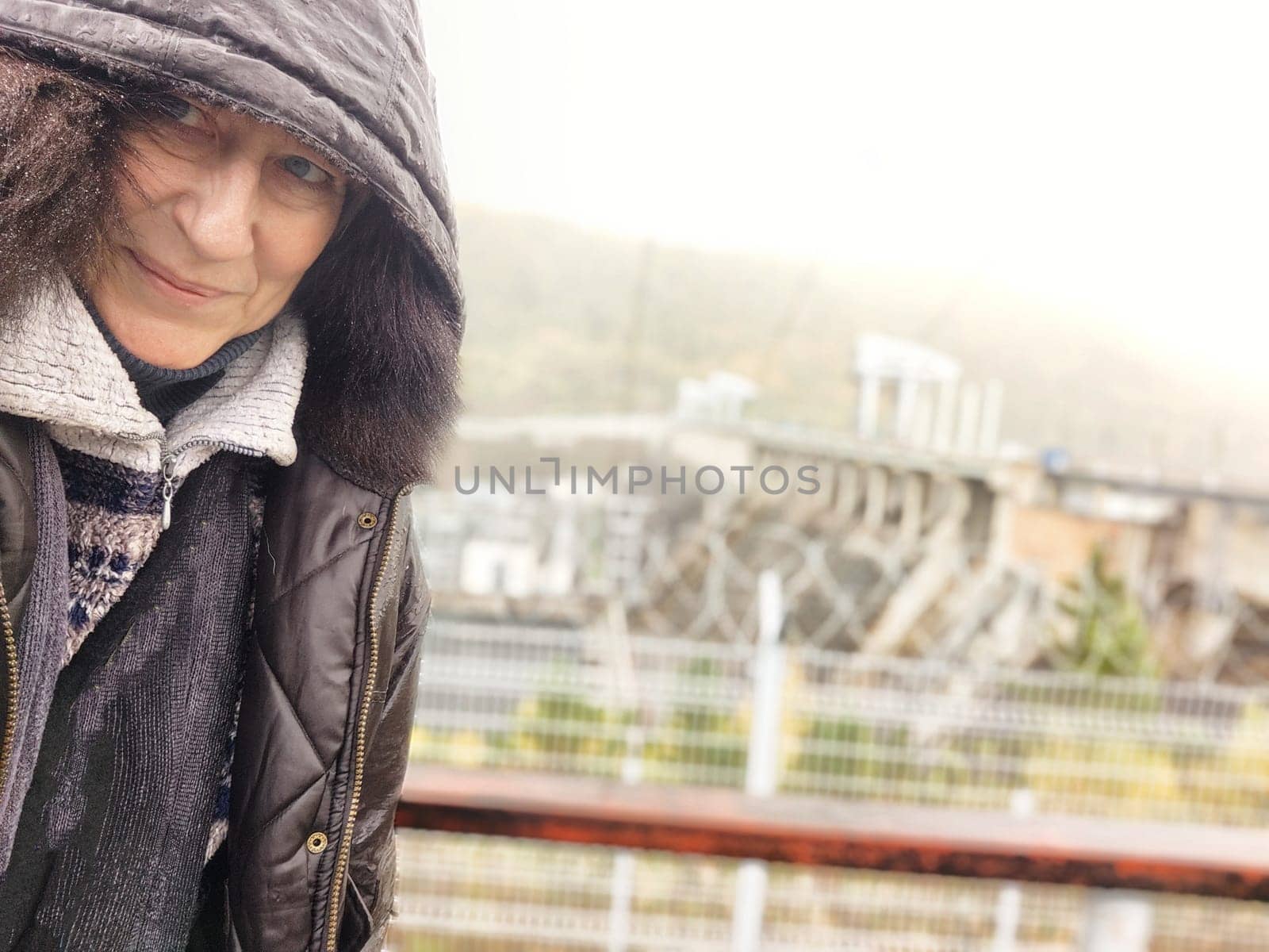 Funny middle-aged adult tourist in jacket during the rain and a hydroelectric power station in the background