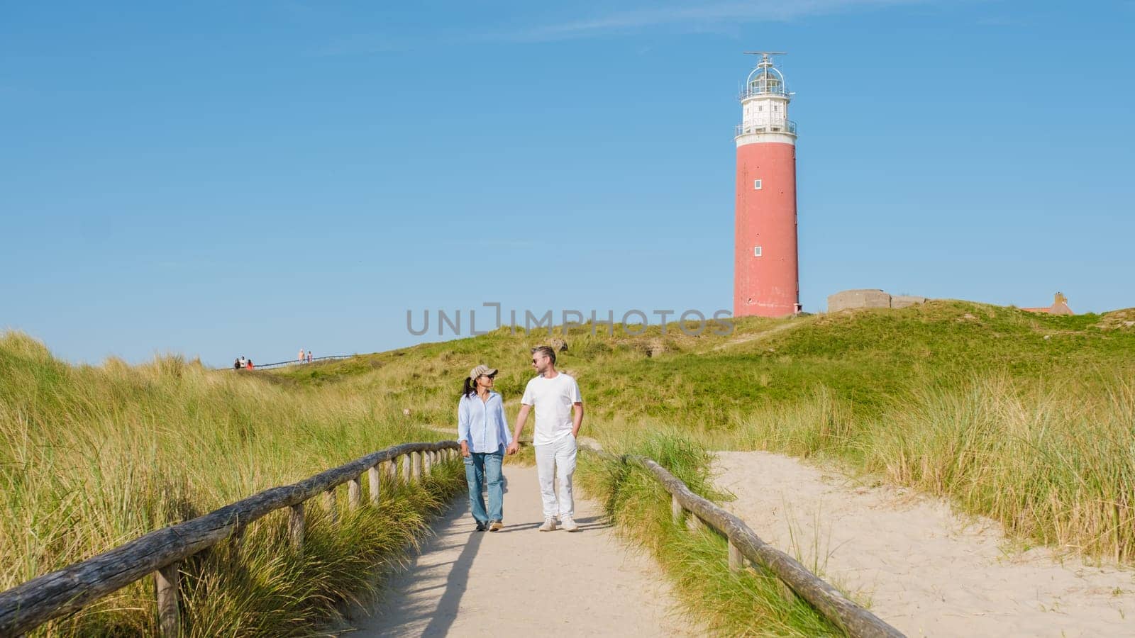 A couple leisurely walks on a path near a picturesque lighthouse in Texel, Netherlands, surrounded by scenic views of the coast. man and woman at The iconic red lighthouse of Texel Netherlands