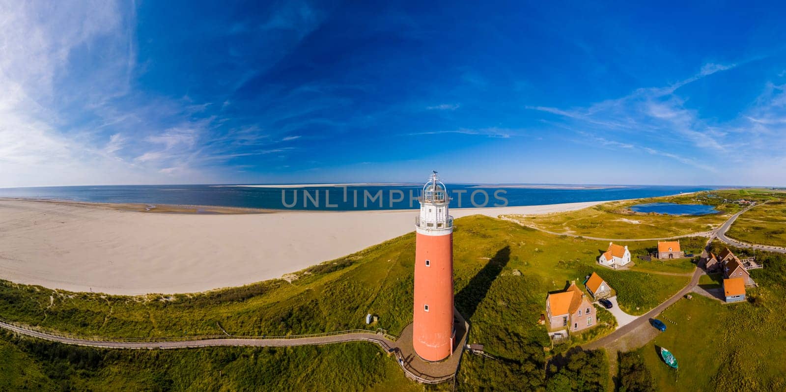 An aerial perspective of a lighthouse standing tall on the sandy beach, overlooking the vast expanse of the ocean with its beacon shining brightly. The iconic red lighthouse of Texel Netherlands