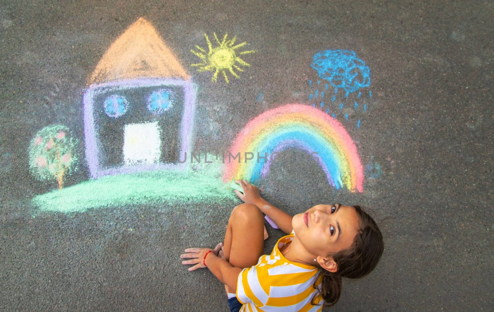 A child draws a house and a rainbow on the asphalt with chalk. Selective focus. Kid.