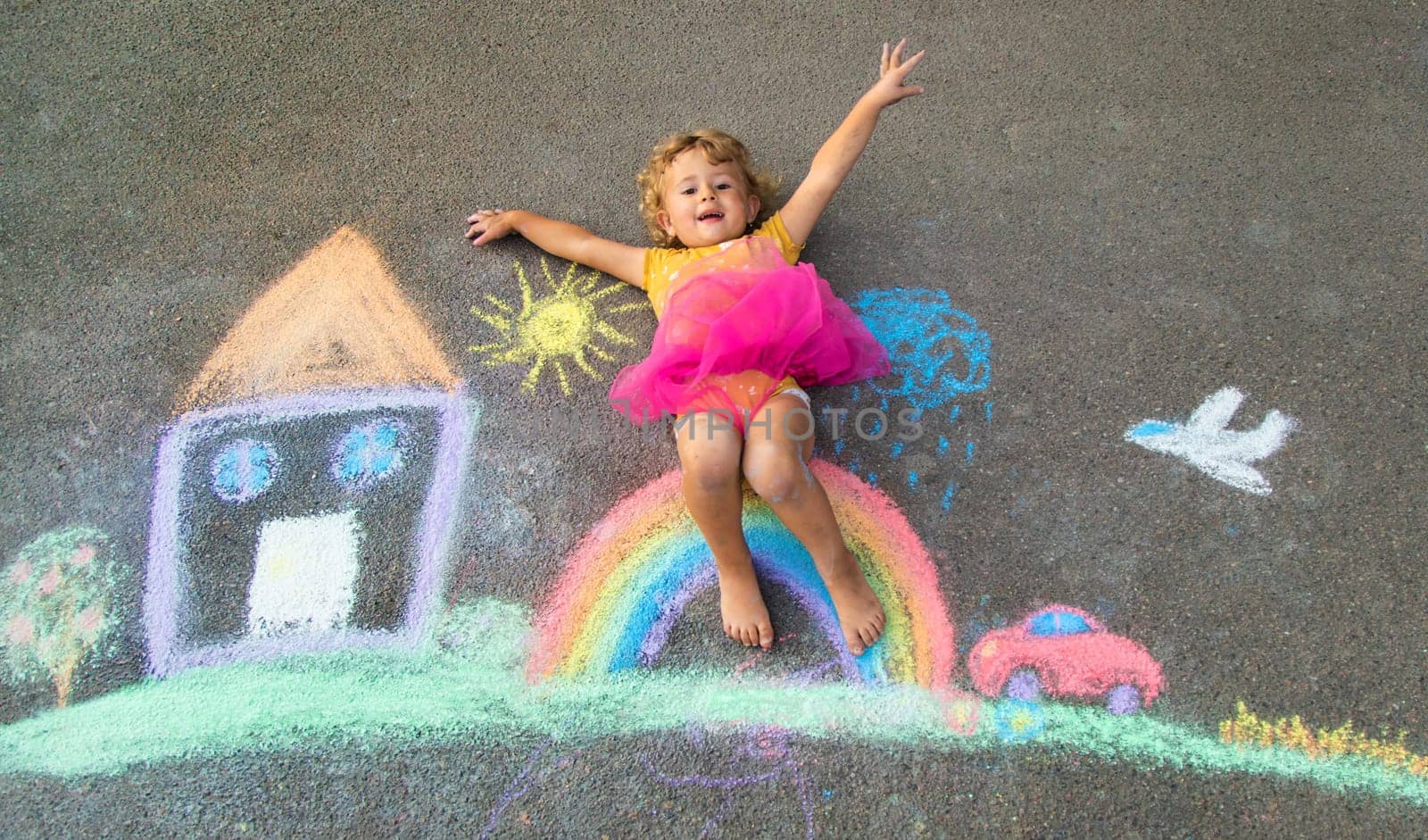 A child draws a house and a rainbow on the asphalt with chalk. Selective focus. Kid.