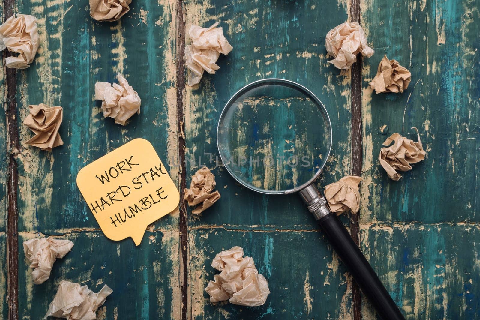 A yellow sticky note with the words work hard, stay humble written on it is placed on top of a pile of crumpled paper. A magnifying glass is also present on the paper, adding a sense of curiosity