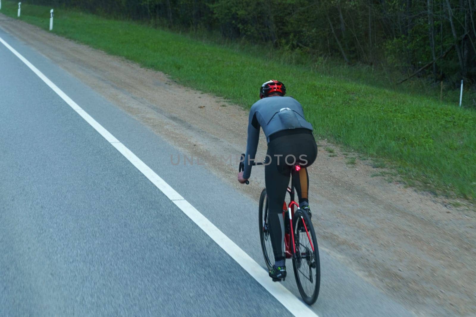Pasvalys, Lithuania - May 3, 2023: A cyclist in a helmet and sportswear rides a road bike along a rural highway with trees in the background, signaling the end of the day.