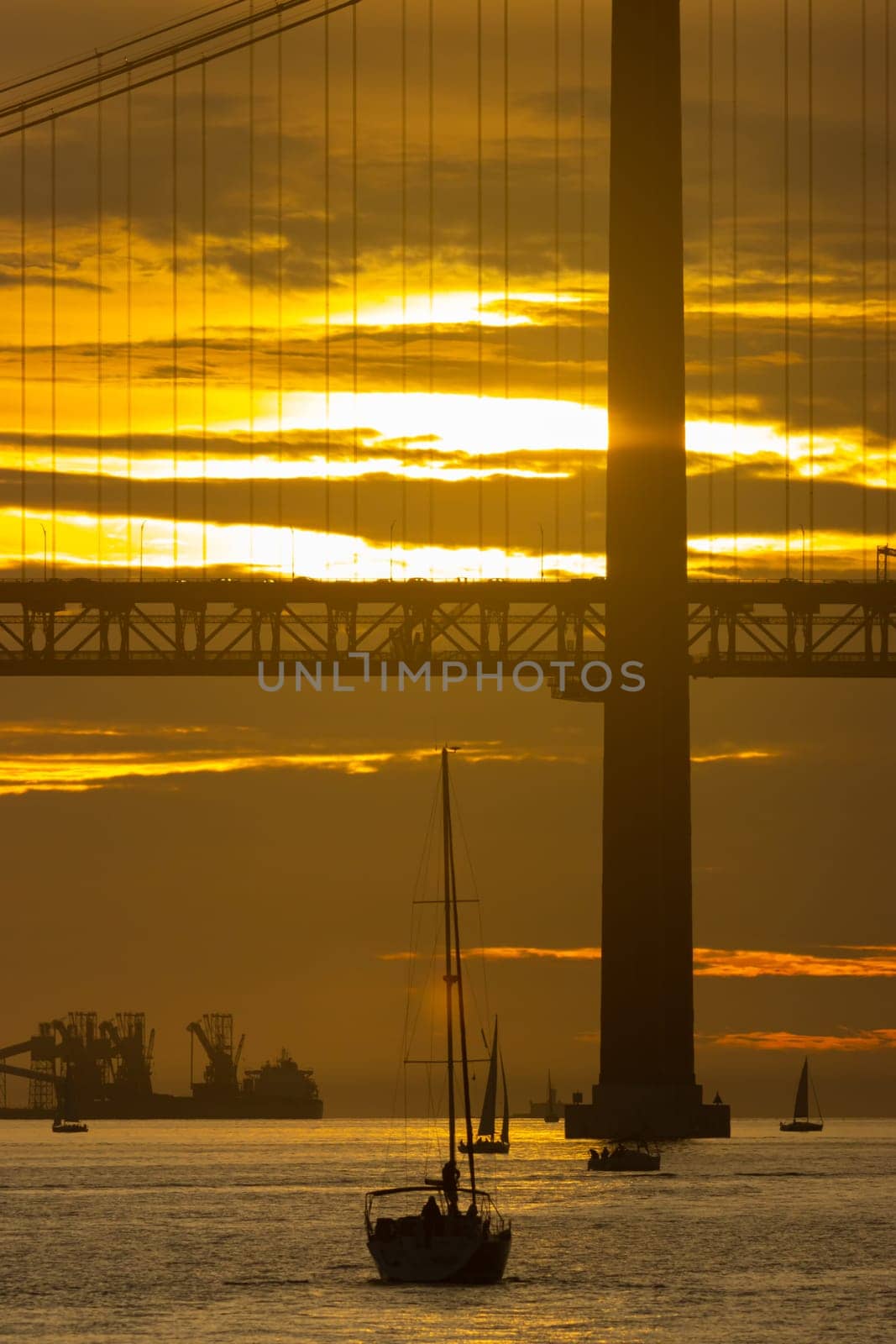 Sailing boat is sailing towards the bridge at bright yellow sunset - oil producing vessel on the background by Studia72