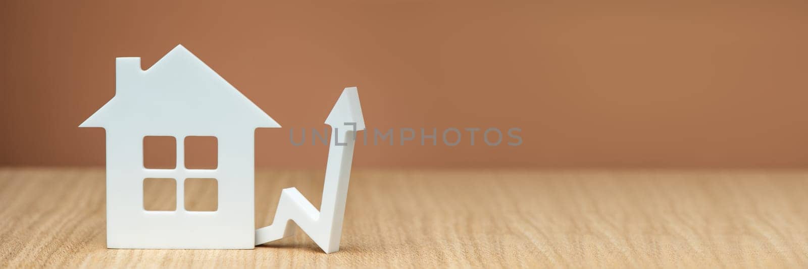 Rising property values. Increasing the cost of housing. Banner with white house model and graphic arrow pointing up close up on brown background. copy space. High quality photo