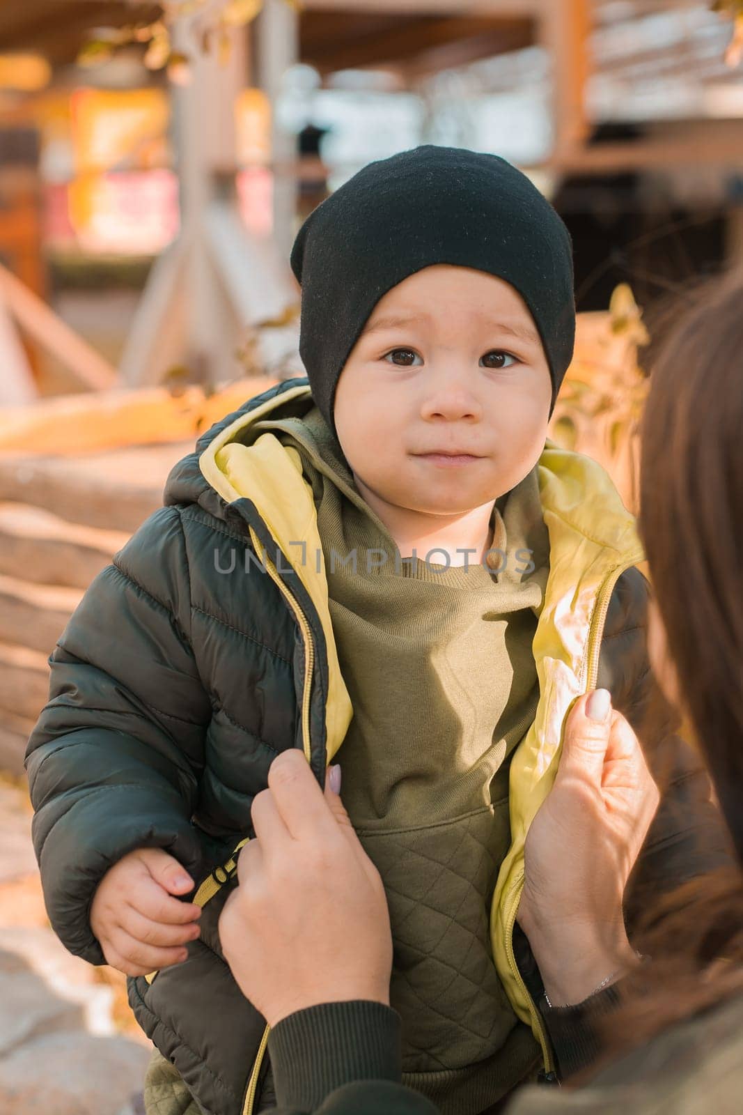 Mother with her little asian son in autumn park close-up. Woman zips up child jacket. Fall and motherhood concept by Satura86