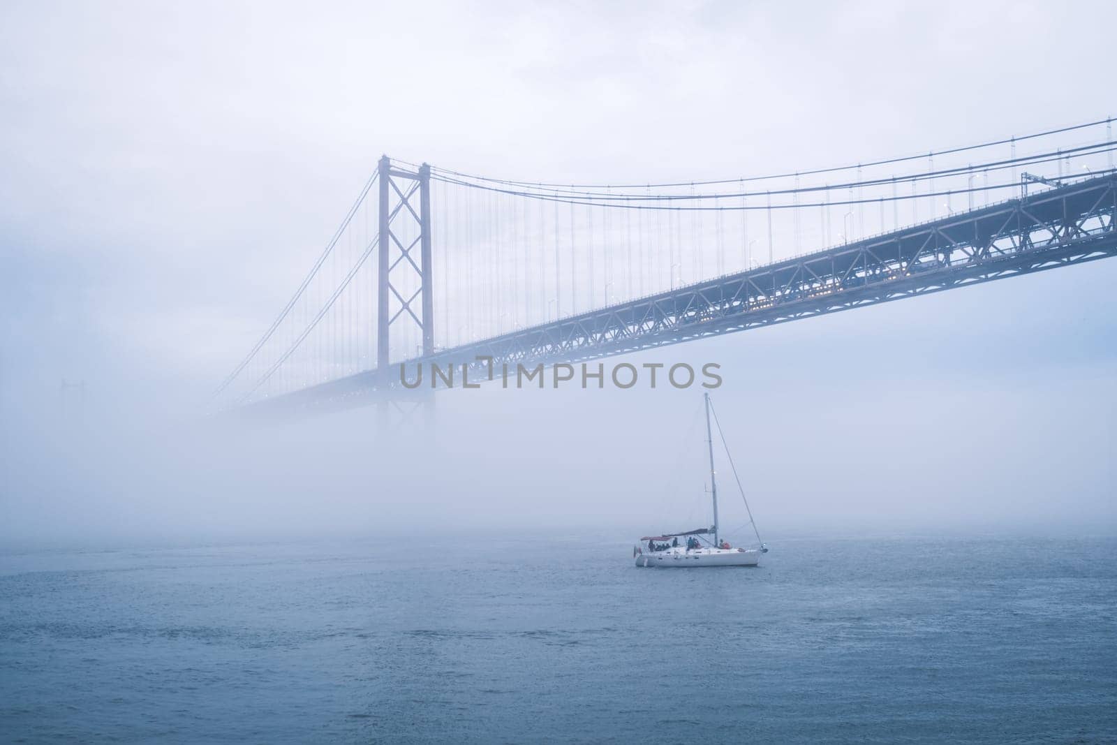 View of 25 de Abril Bridge famous tourist landmark of Lisbon connecting Lisboa and Almada in heavy fog mist wtih yacht boats passing under. Lisbon, Portugal