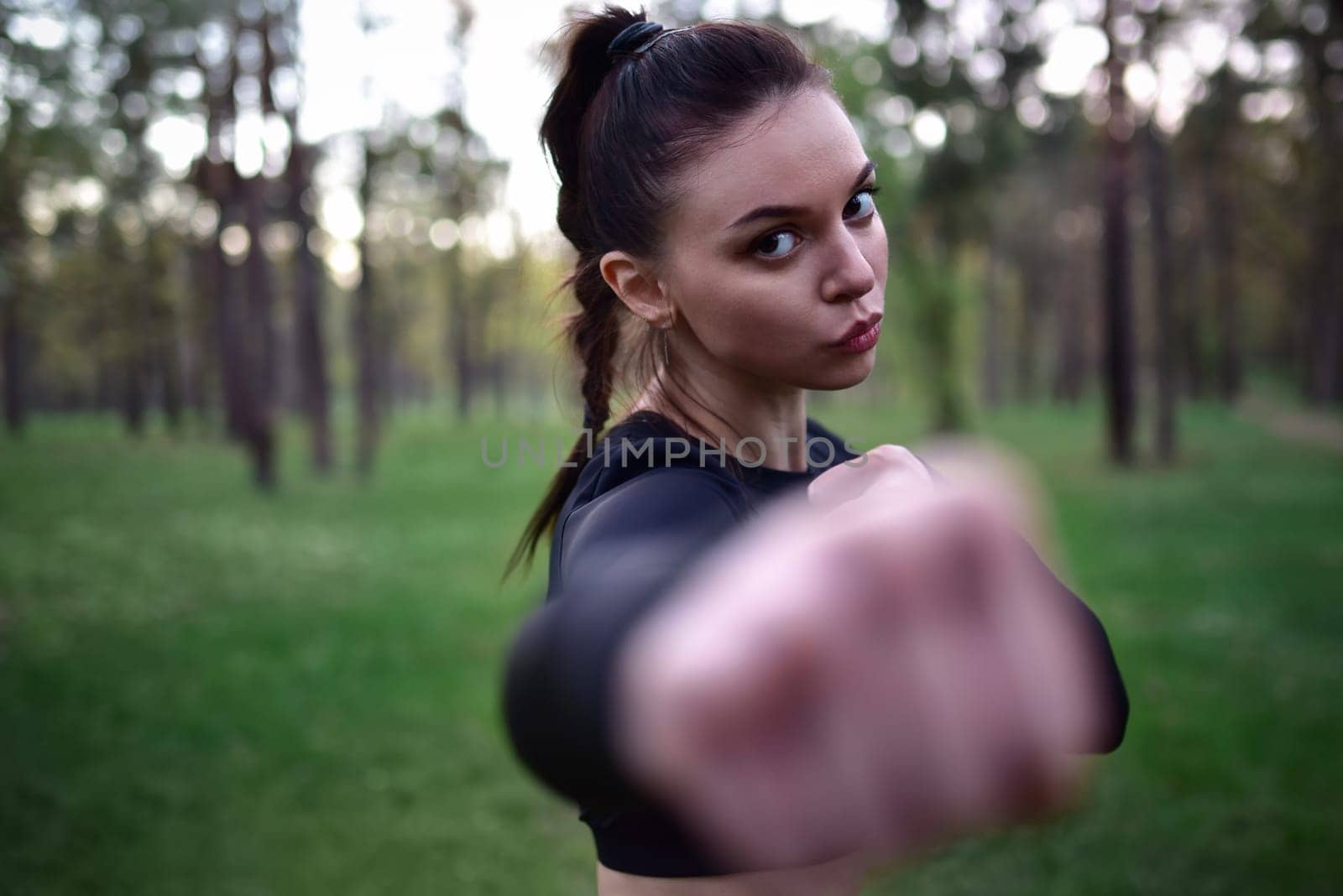 Young woman doing martial arts training in sporty black top holding hands in fighting pose against forest background.