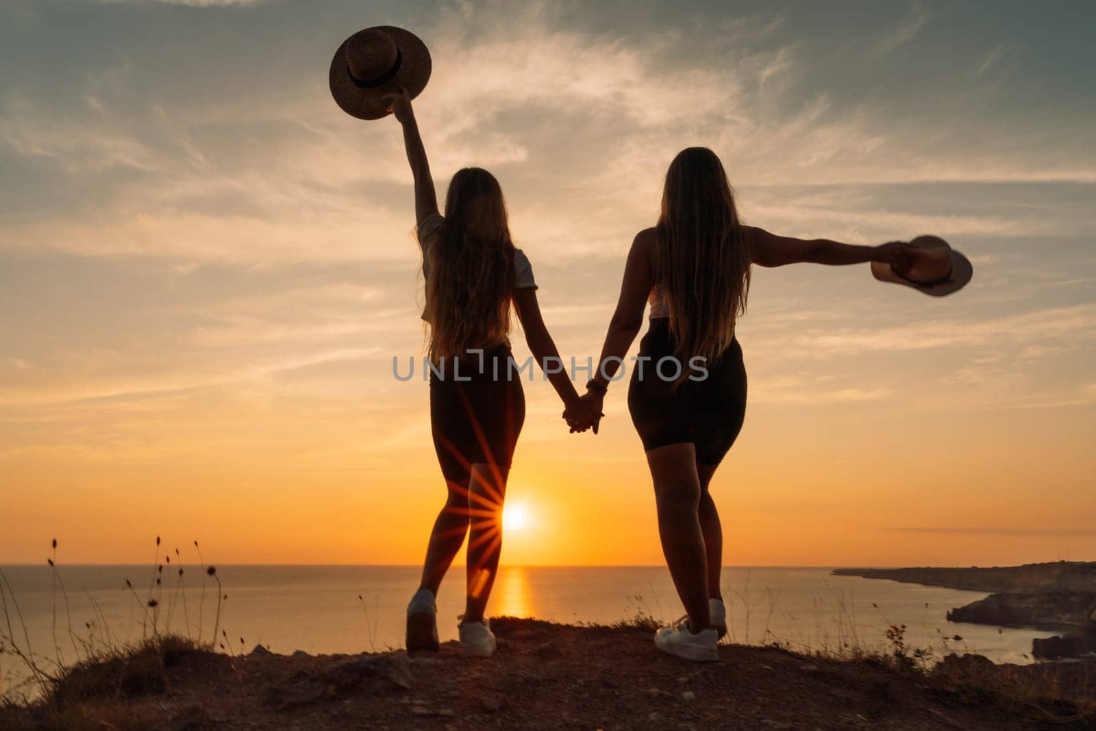 Two women are standing on a hillside, holding hands and wearing hats. The sun is setting in the background, creating a warm and peaceful atmosphere
