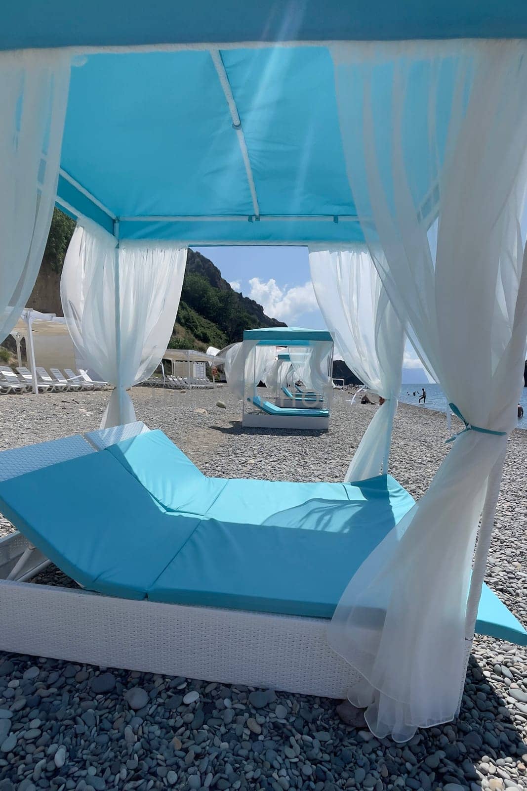A blue and white beach umbrella with white curtains is set up on a beach. The umbrella is open and the white curtains are spread out, creating a relaxing and inviting atmosphere. The beach is empty